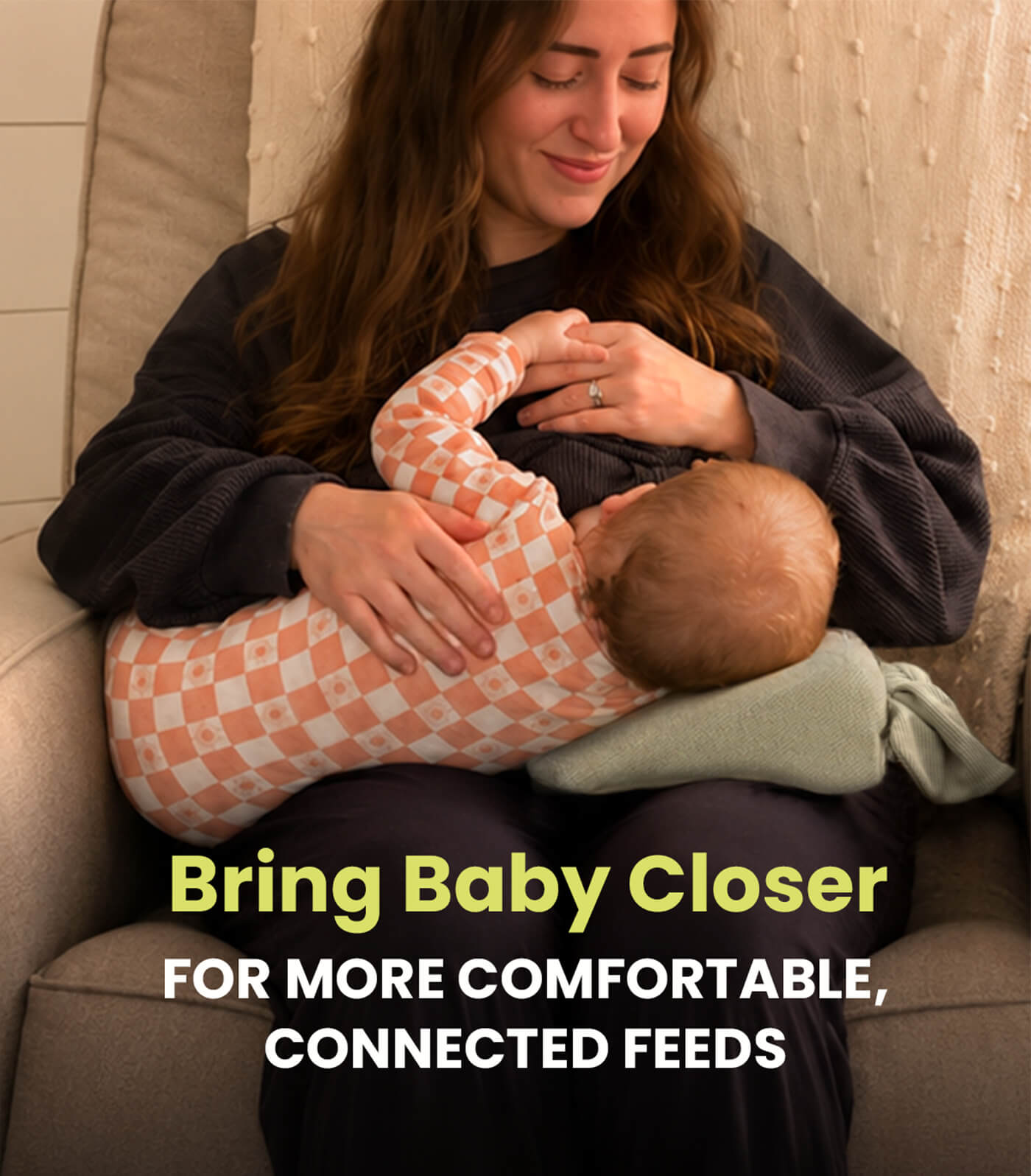 A woman in a dark sweatshirt holds a baby wearing a peach and white checkered outfit while breastfeeding. The image features a text overlay at the bottom that reads 'Bring Baby Closer FOR MORE COMFORTABLE, CONNECTED FEEDS'. The woman is using the acute baby co wedge nursing pillow to comfortably feed her baby.